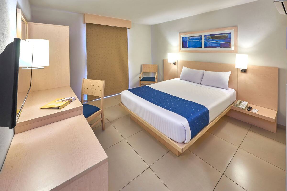 A hotel room with a bed, a desk with a chair, a telephone, and a wall-mounted TV.