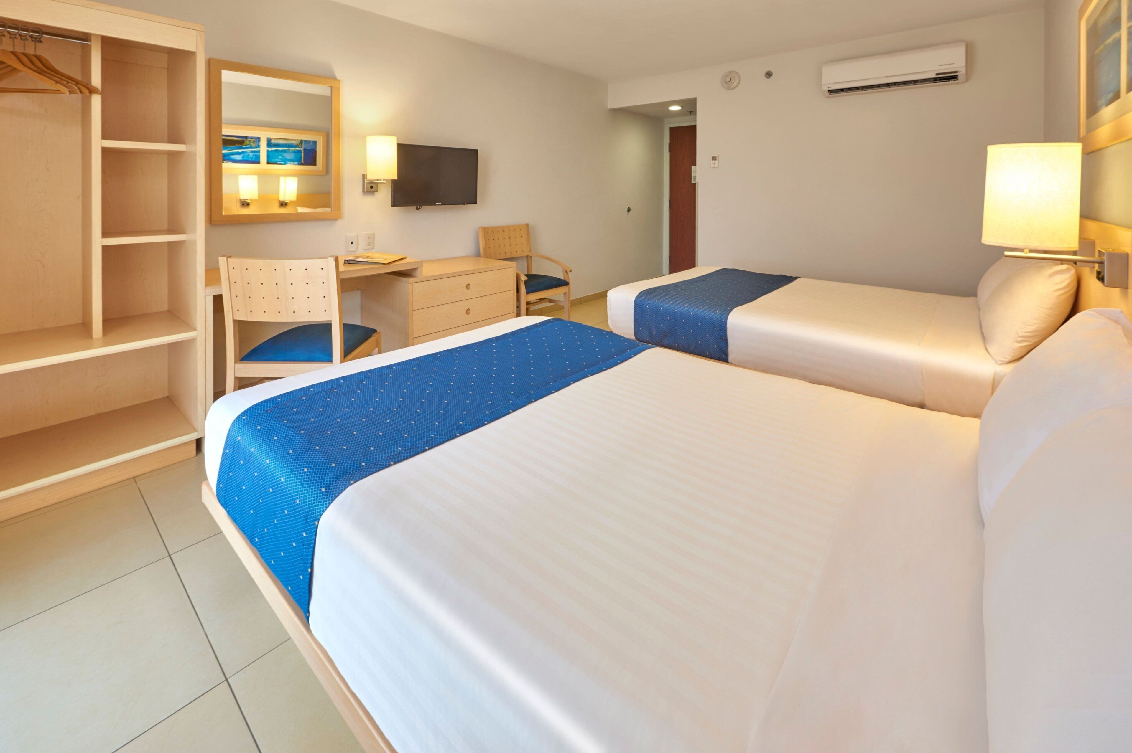 Une chambre d’hôtel avec deux lits, un bureau, une chaise, une armoire, une télévision et un appareil de climatisation.