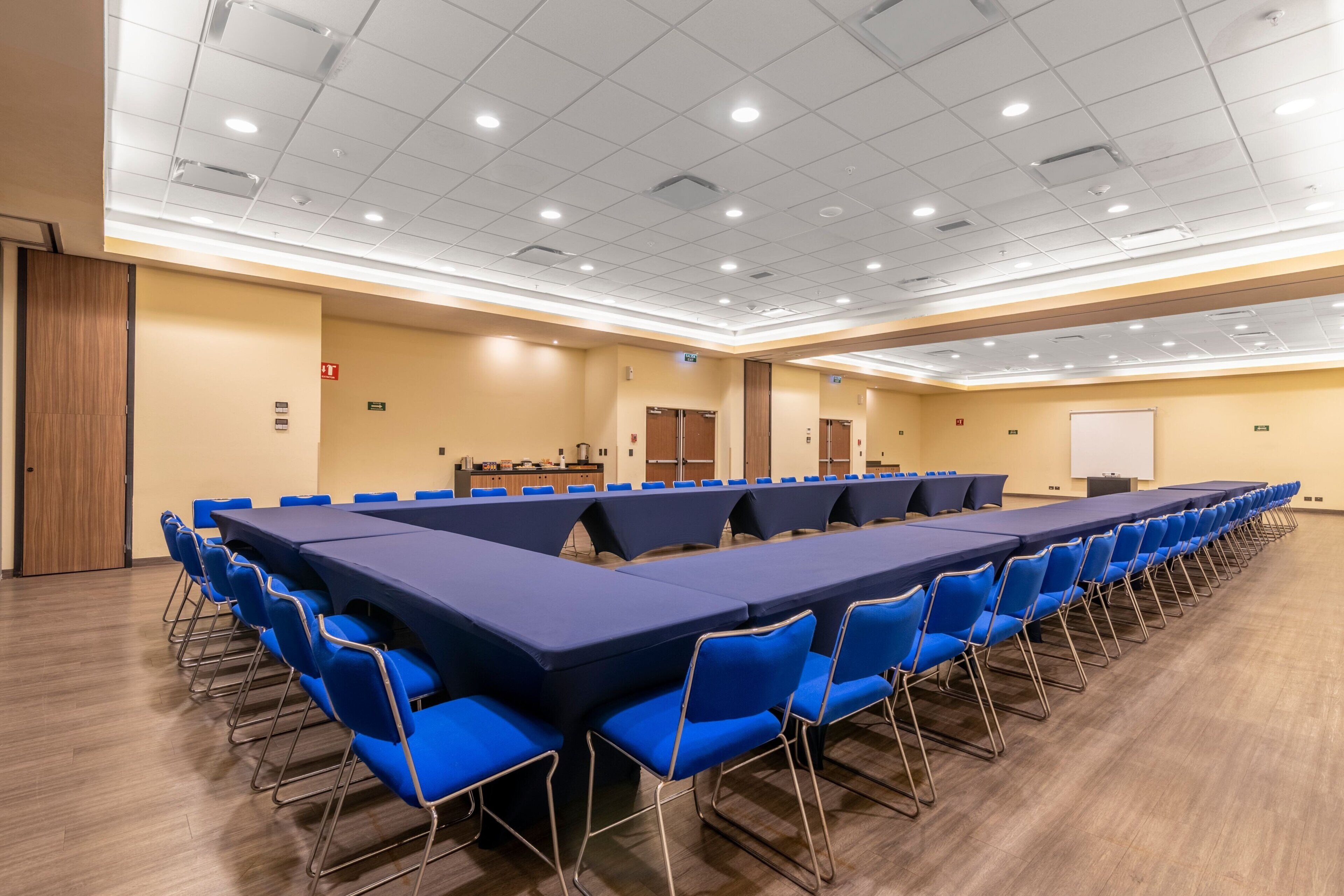 Uma sala de conferência com uma mesa comprida e cadeiras azuis, equipada com um quadro branco e um púlpito.