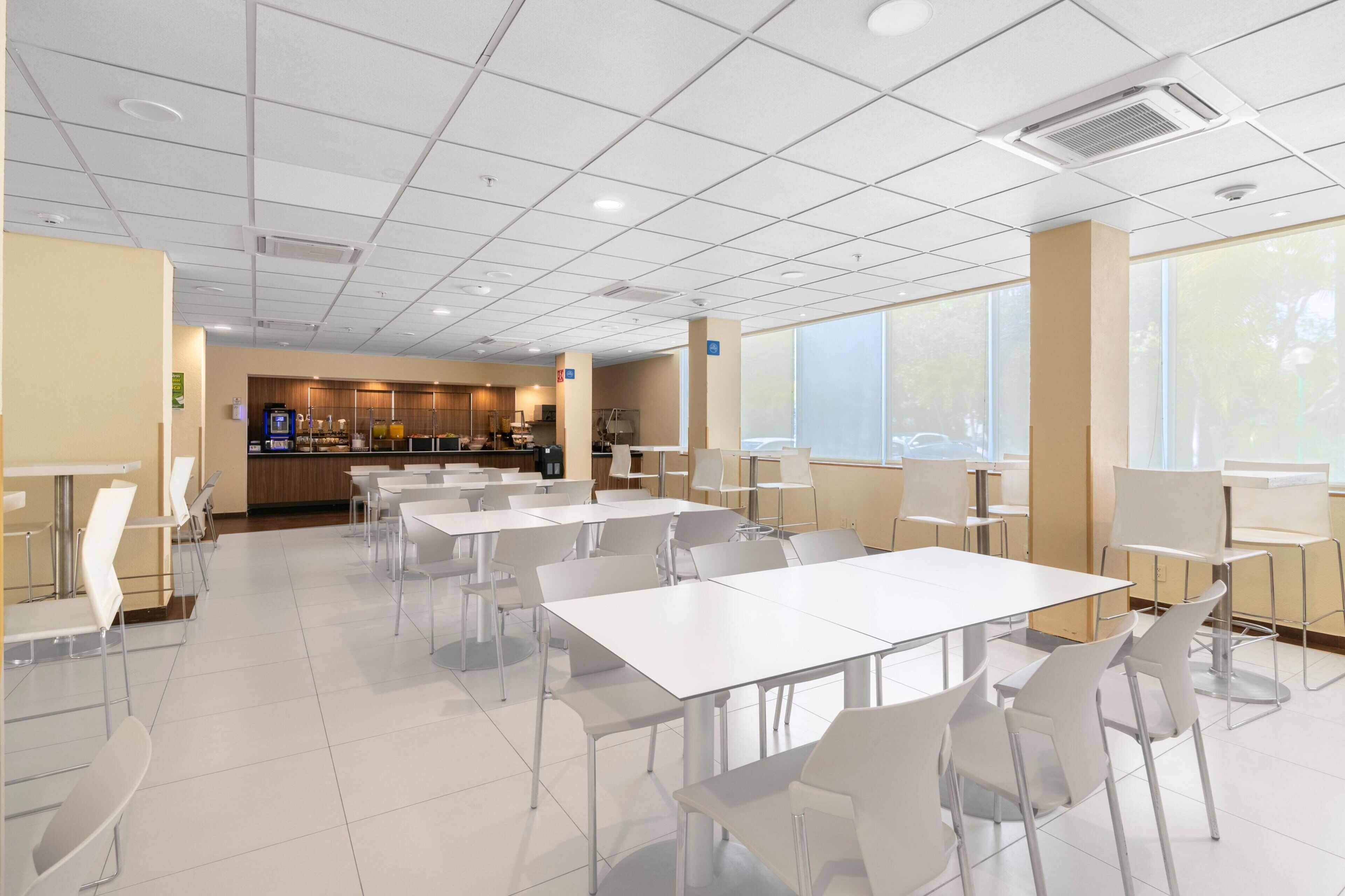 Een ruime cafetaria met witte tafels en stoelen, een keuken in de achtergrond, en grote ramen die natuurlijk licht binnenlaten.