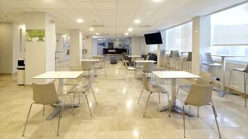 A spacious indoor area with multiple tables and chairs arranged in rows, a reception desk, and a large screen mounted on the wall.