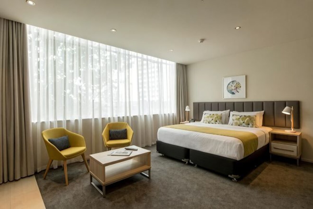 A hotel room with a large bed, two chairs, a bedside table, and a window with curtains.