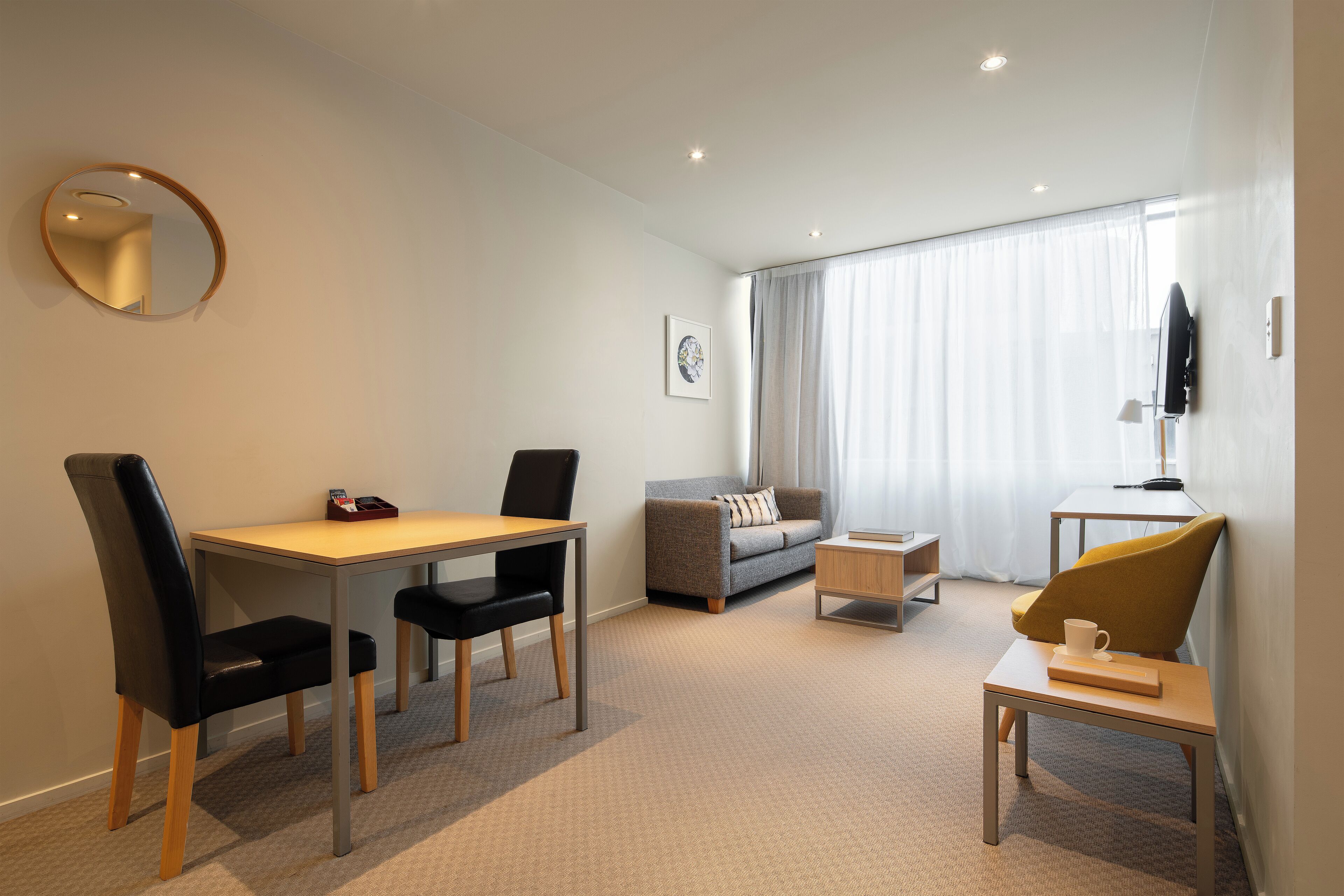 A modern hotel room with a dining area, a sofa, a TV, and a window with curtains.