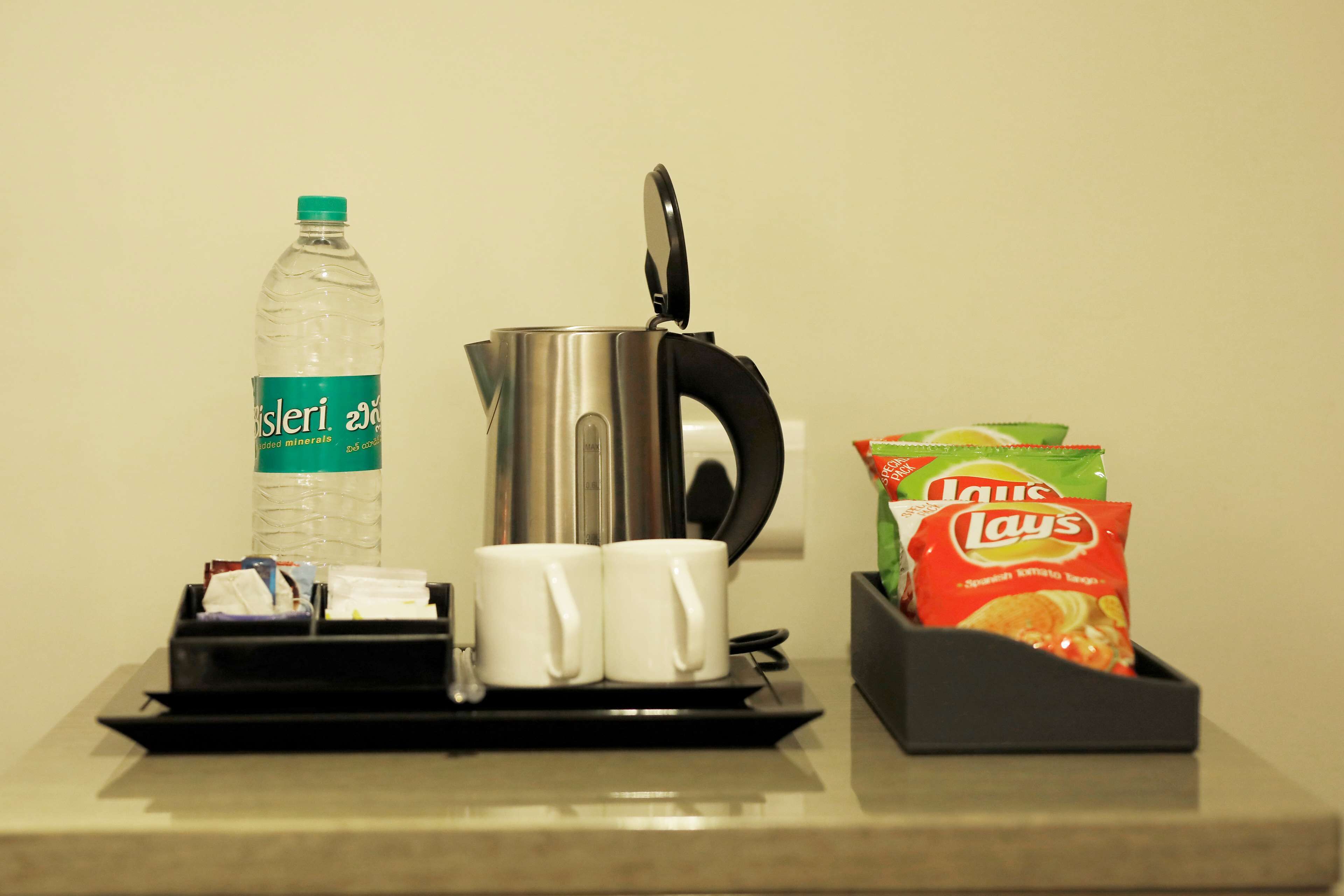 A kitchen counter with a kettle, two white mugs, a bottle of water, and a bag of Lay's potato chips.