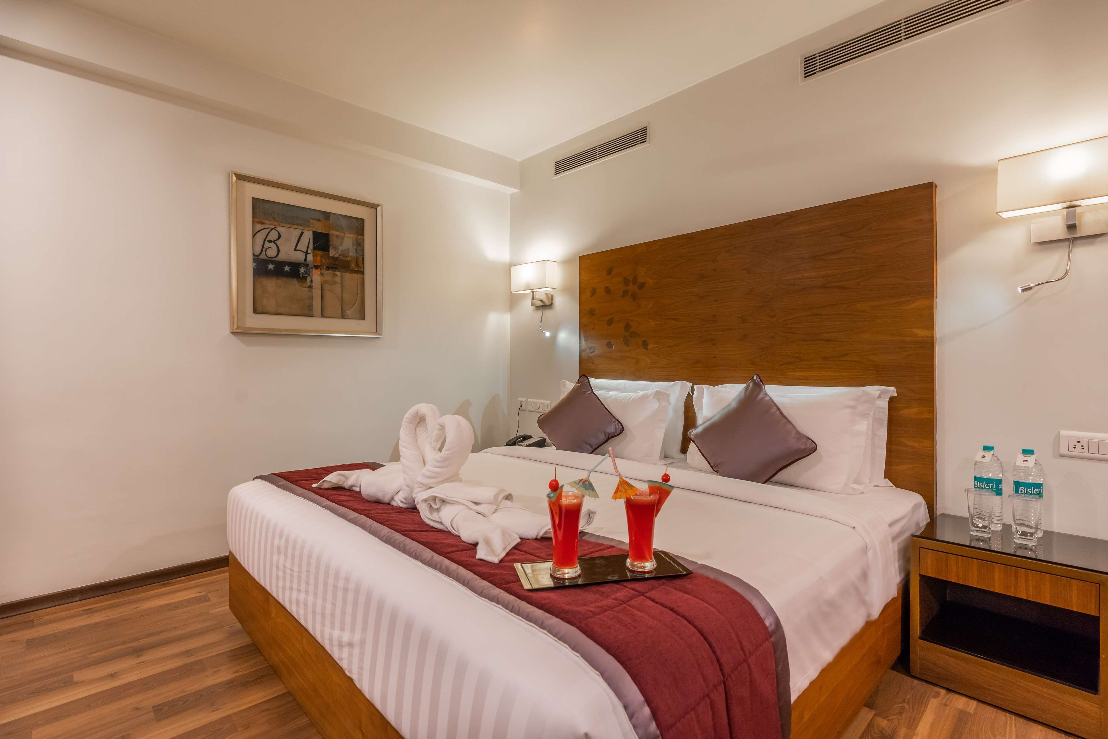 Ein Hotelzimmer mit einem hölzernen Kopfteil, einem ordentlich bezogenen Bett mit roter Decke, zwei Wasserflaschen und einem gerahmten Bild an der Wand.