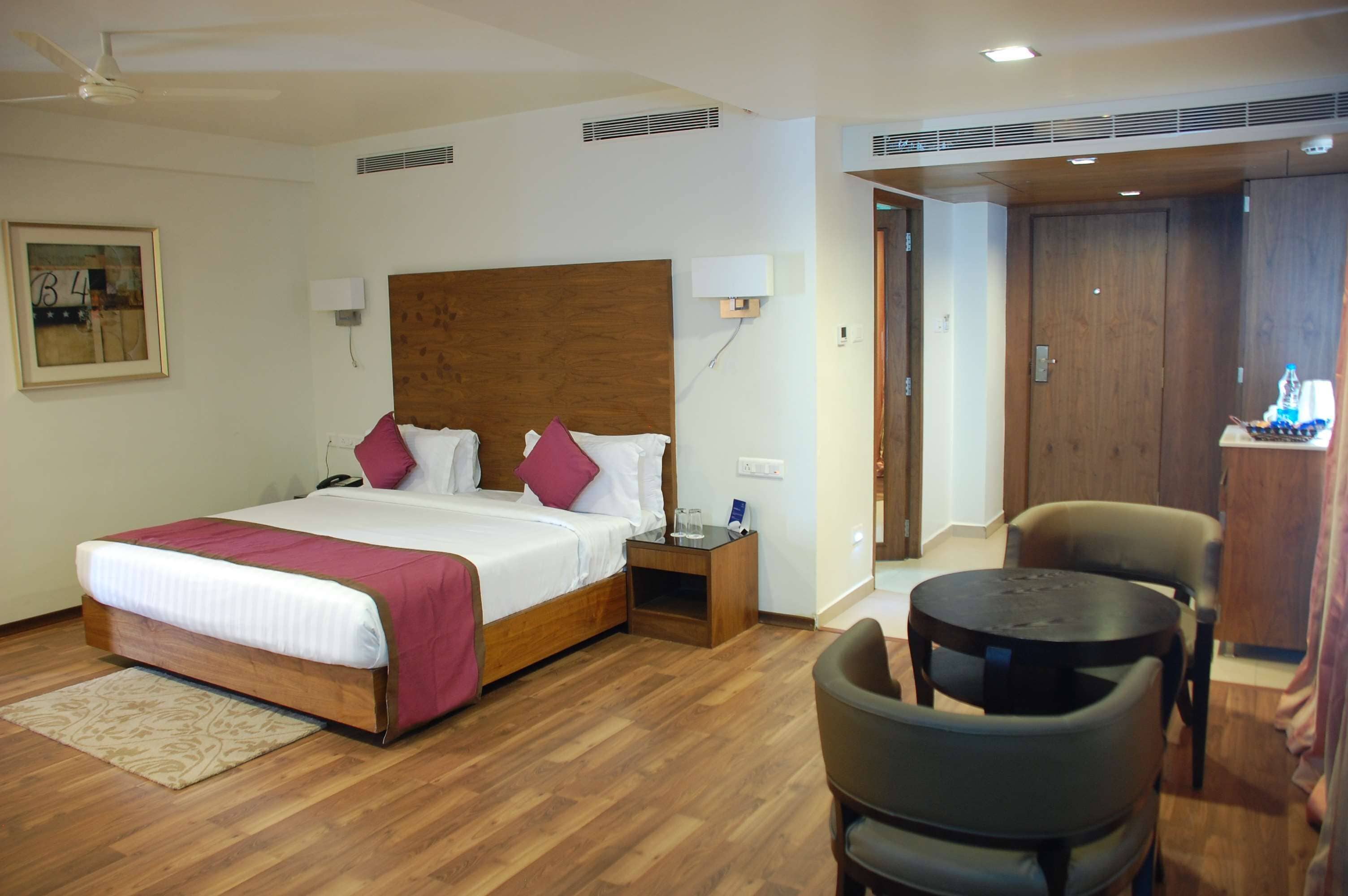 Habitación de hotel con cama de madera, una mesita de noche, una silla, una mesa redonda pequeña y una puerta que da a otra habitación.