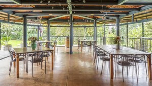 Un pabellón al aire libre con varias mesas y sillas, ventanas amplias y vistas a la vegetación circundante.