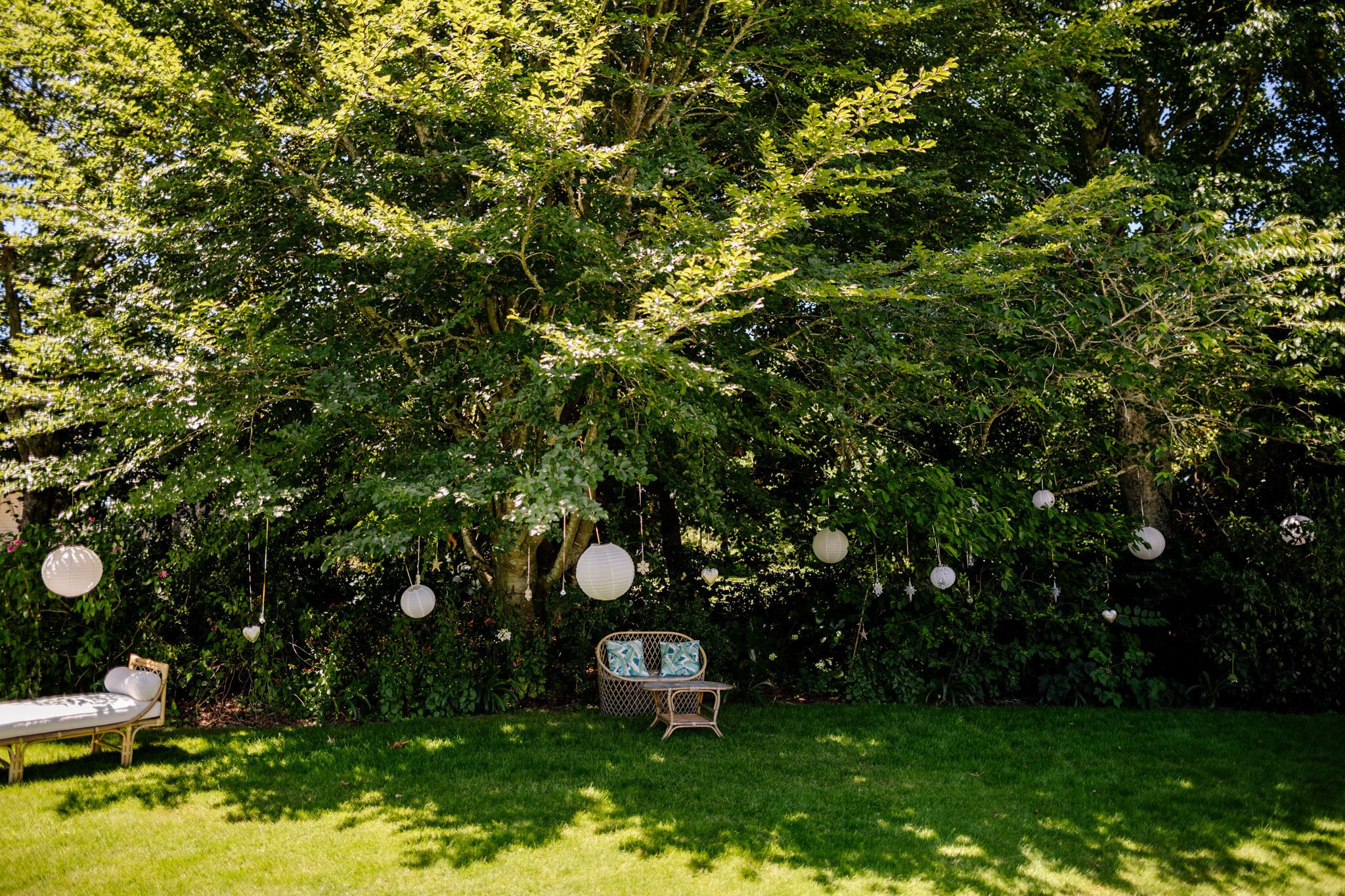 A garden with a wicker sofa and a dining table, surrounded by lush greenery and hanging paper lanterns.