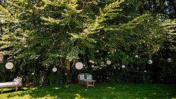 A garden with a wicker sofa and a dining table, surrounded by lush greenery and hanging paper lanterns.