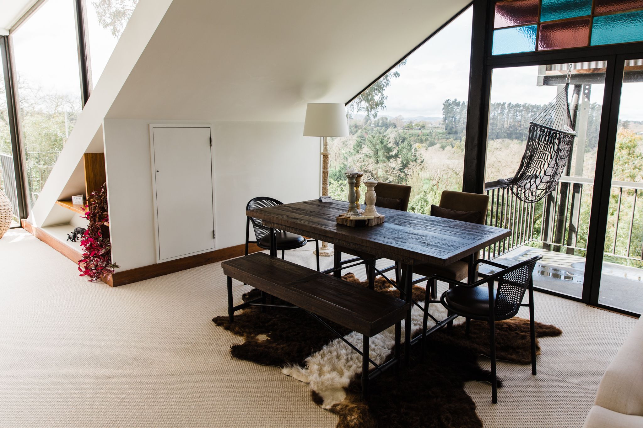 Een eethoek met een houten tafel, stoelen en een bank. Op de tafel staat een lamp, en er is uitzicht op het buitenlands landschap door grote ramen.