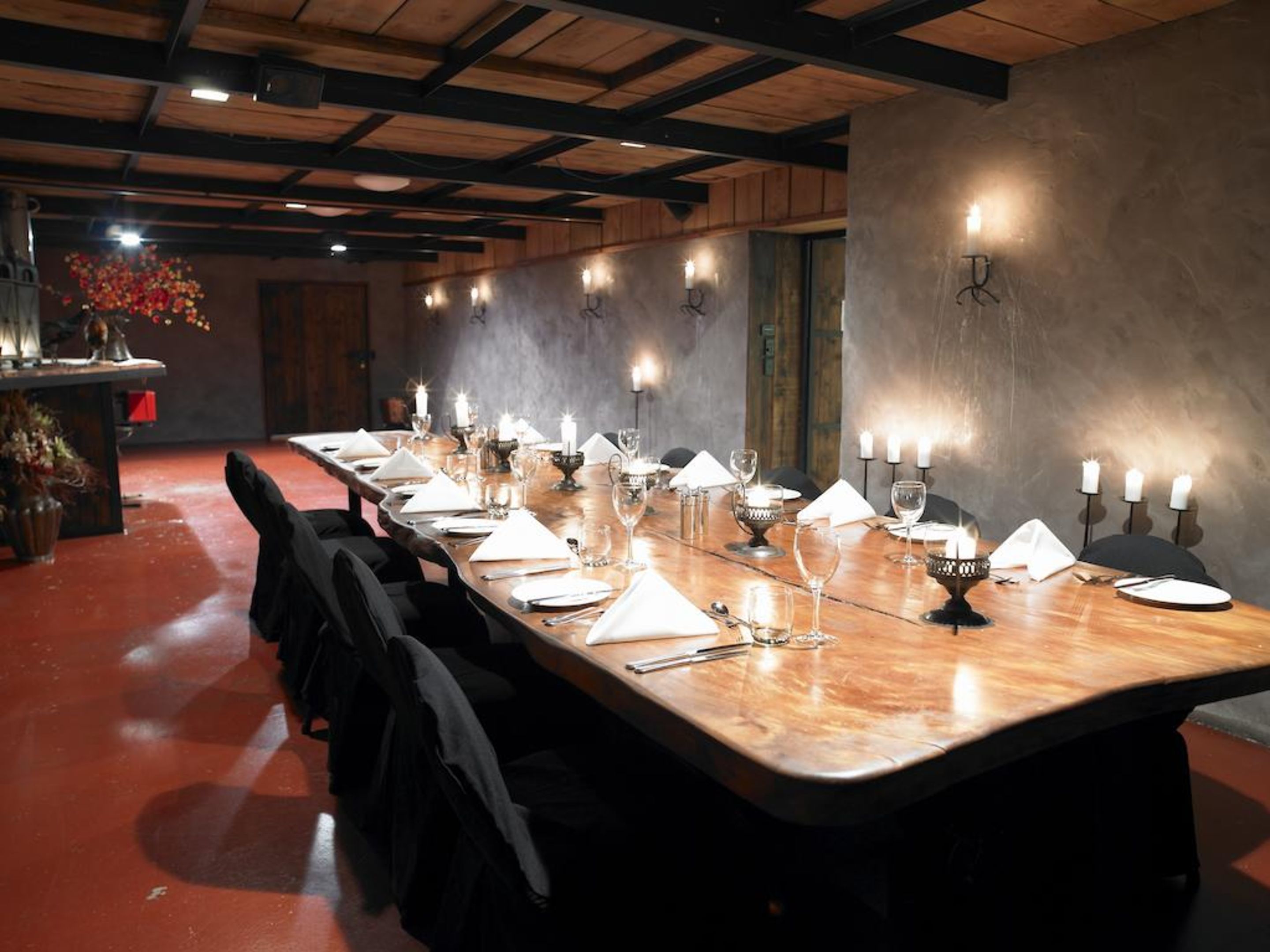 Una larga mesa de comedor preparada para una comida, con velas y cristalería, en una habitación con vigas de madera y pared de hormigón.