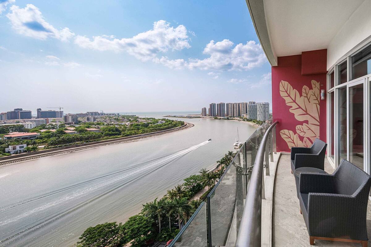 A balcony with a view of a curved road, a beach, and high-rise buildings in the distance.