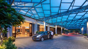 Una entrada de hotel moderna con techo de vidrio, un coche estacionado afuera y una persona abriendo la puerta del vehículo.