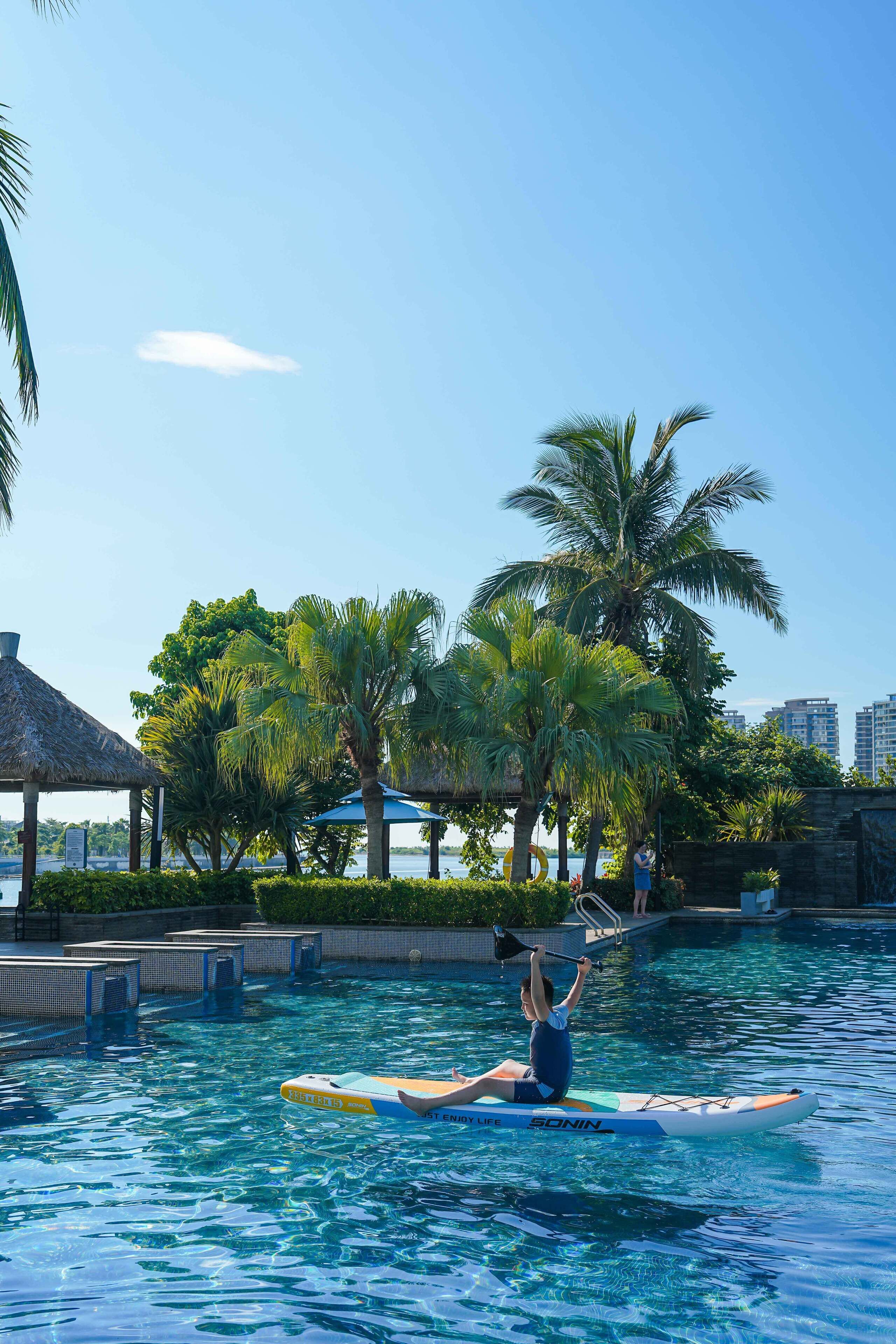 Una persona haciendo paddleboard en una piscina rodeada de palmeras y una estructura con techo de paja.