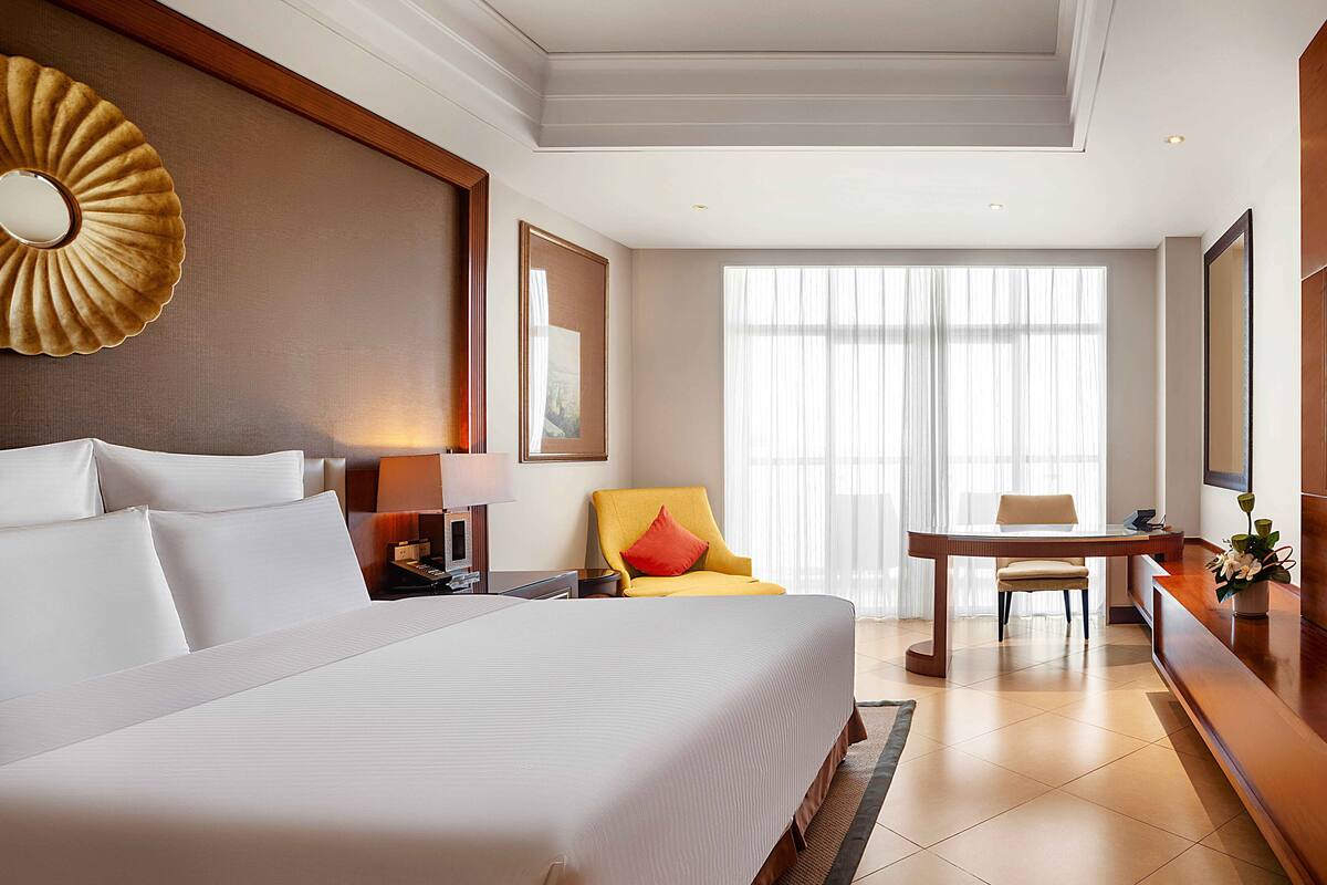 A hotel room with a large bed, a desk, a chair, and a window with sheer curtains.