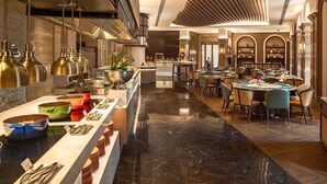 Un moderno vestíbulo de hotel con suelo de mármol, una zona de bar central con diversas opciones de comida y un comedor con mesas y sillas redondas.