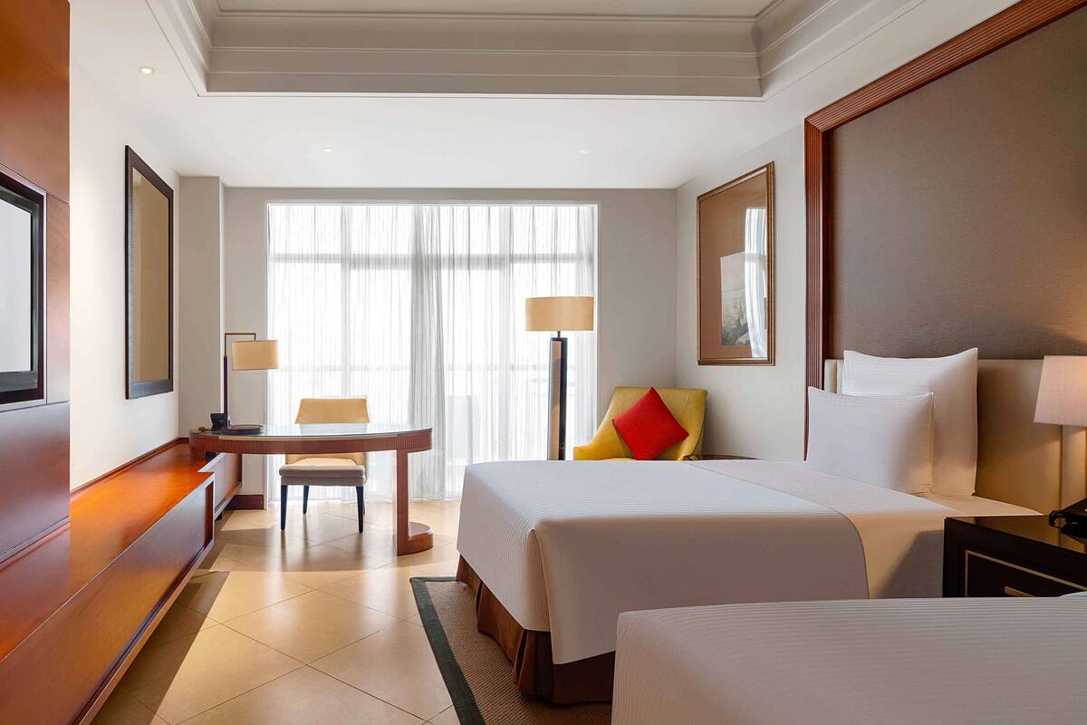 A hotel room with two beds, a desk, a chair, and a large window with sheer curtains.
