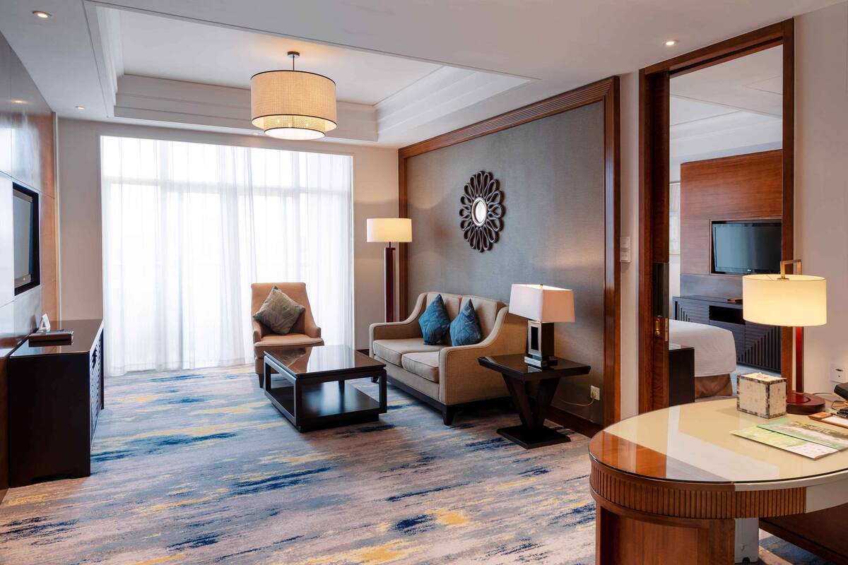 A modern hotel room with a large window, a sofa, a coffee table, a TV, and a desk with a lamp.