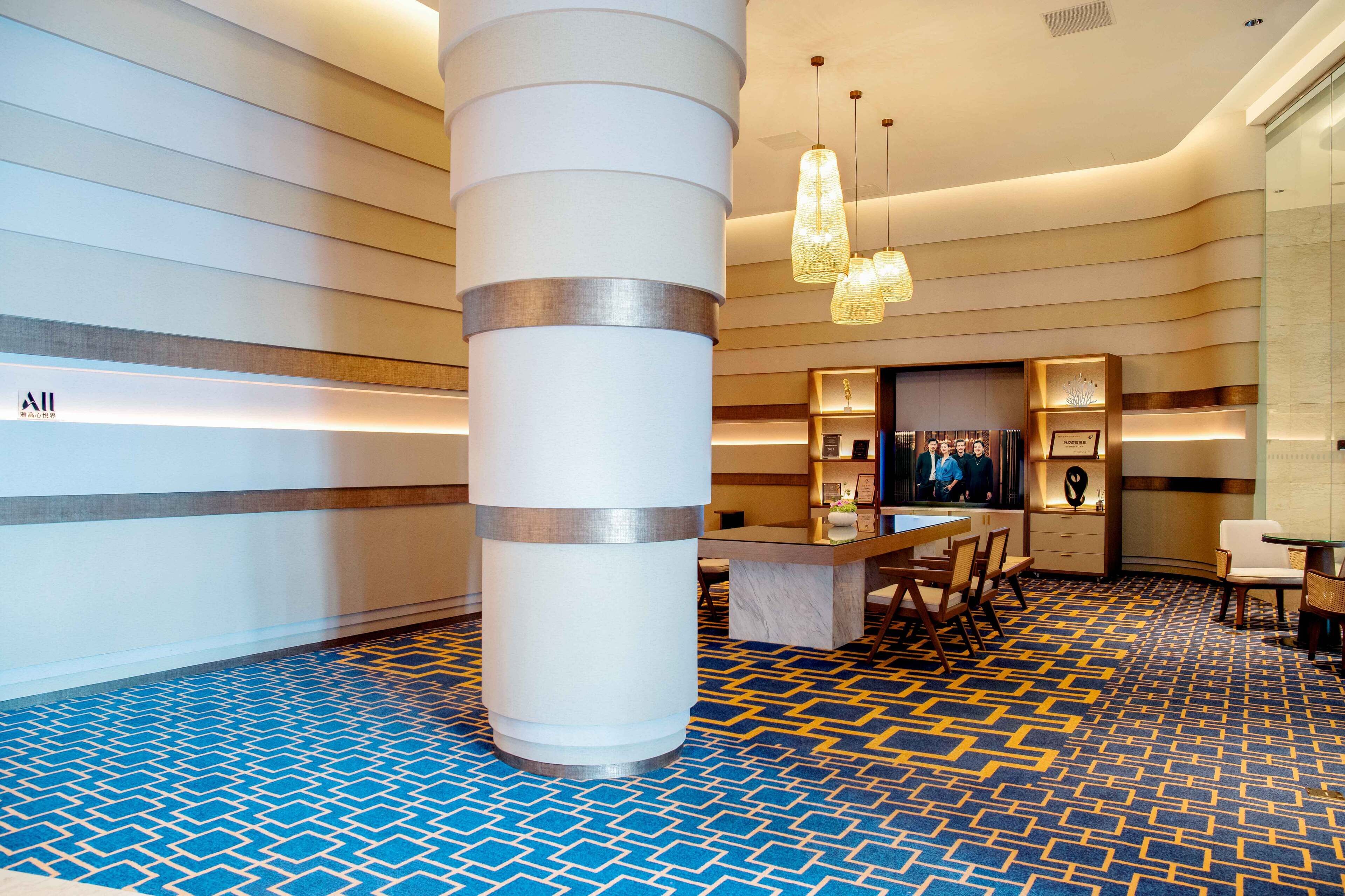 Eine Hotelhalle mit einem gemusterten Teppich in Blau und Gelb, einer zentralen Säule, einem Empfangstresen und Pendelleuchten.
