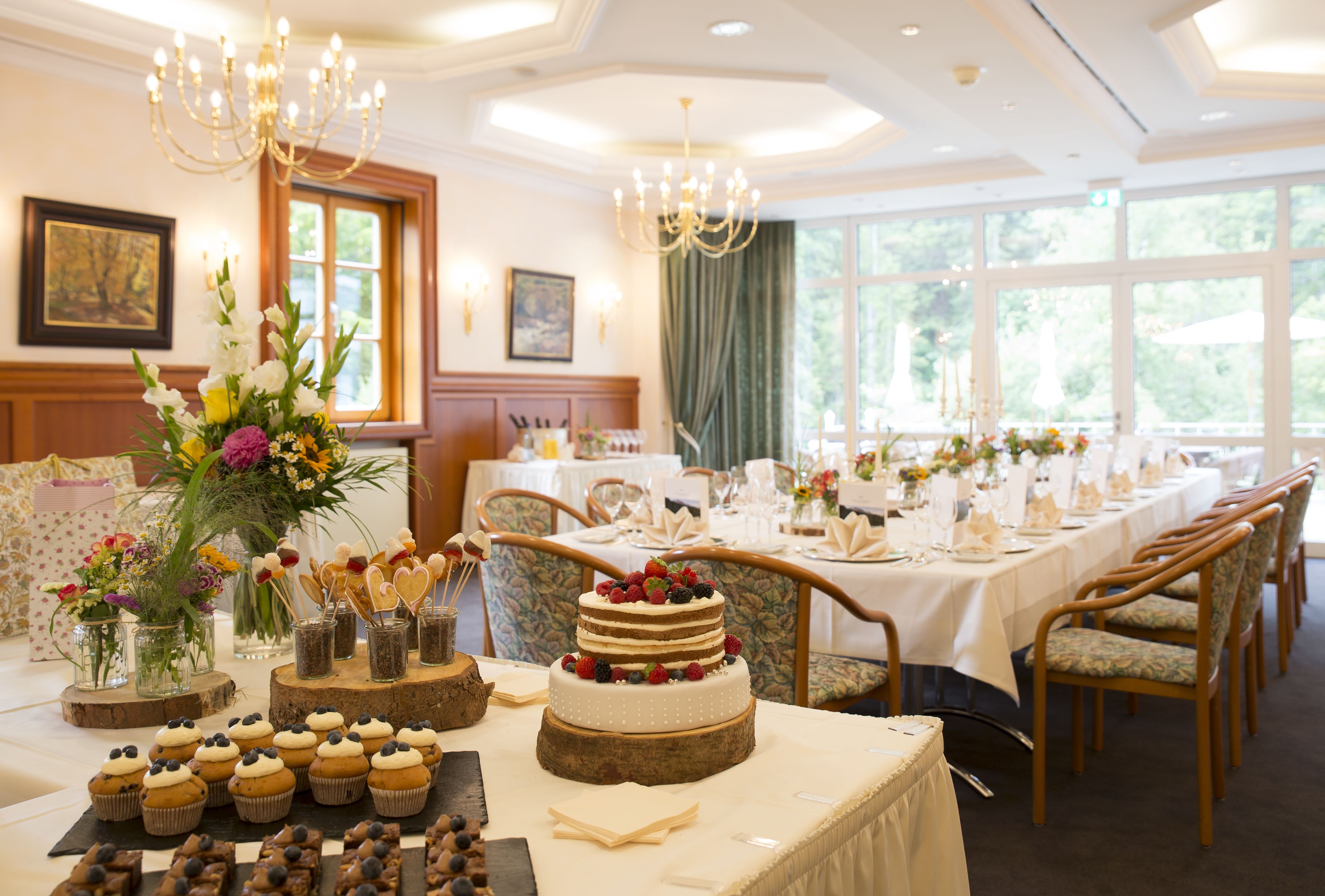 Une salle de banquet avec une longue table dressée pour un événement formel, agrémentée d’un gâteau à étages, de petits gâteaux et de compositions florales.