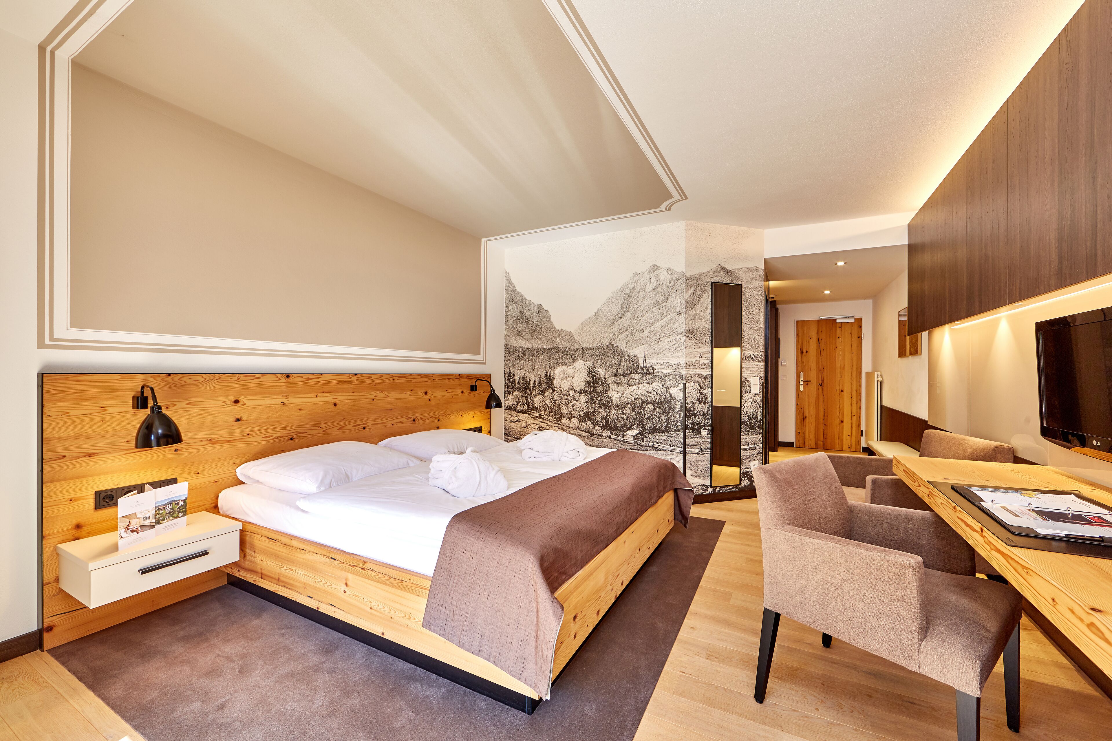 A modern hotel room with a wooden bed, a desk with a chair, a large mirror, and a mountain view mural.