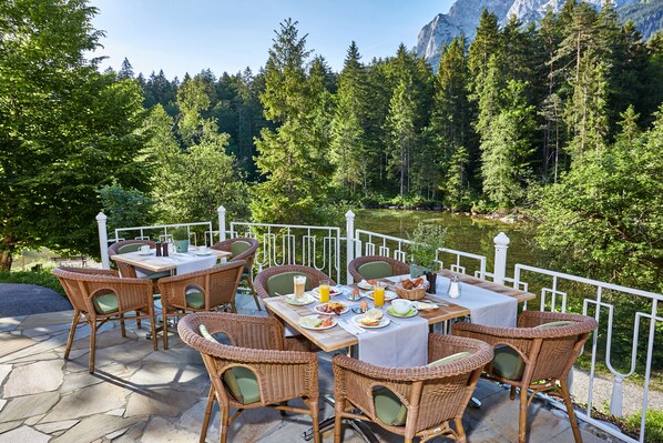 An outdoor dining area with wicker chairs and tables, set against a backdrop of a lake and forested mountains. - Hotel am Badersee (Grainau)