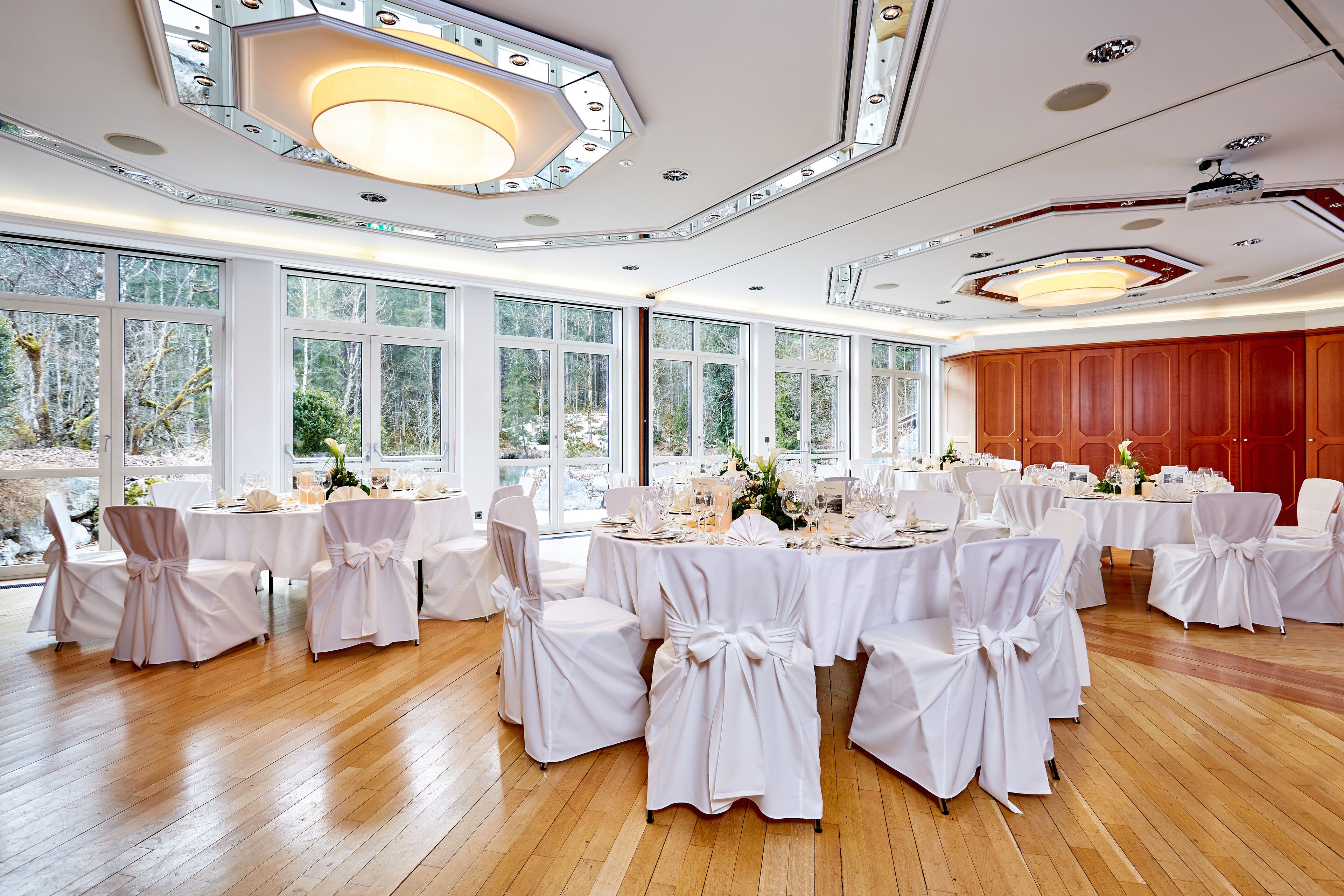 A banquet hall with round tables set for a formal event, large windows, and a wooden floor.