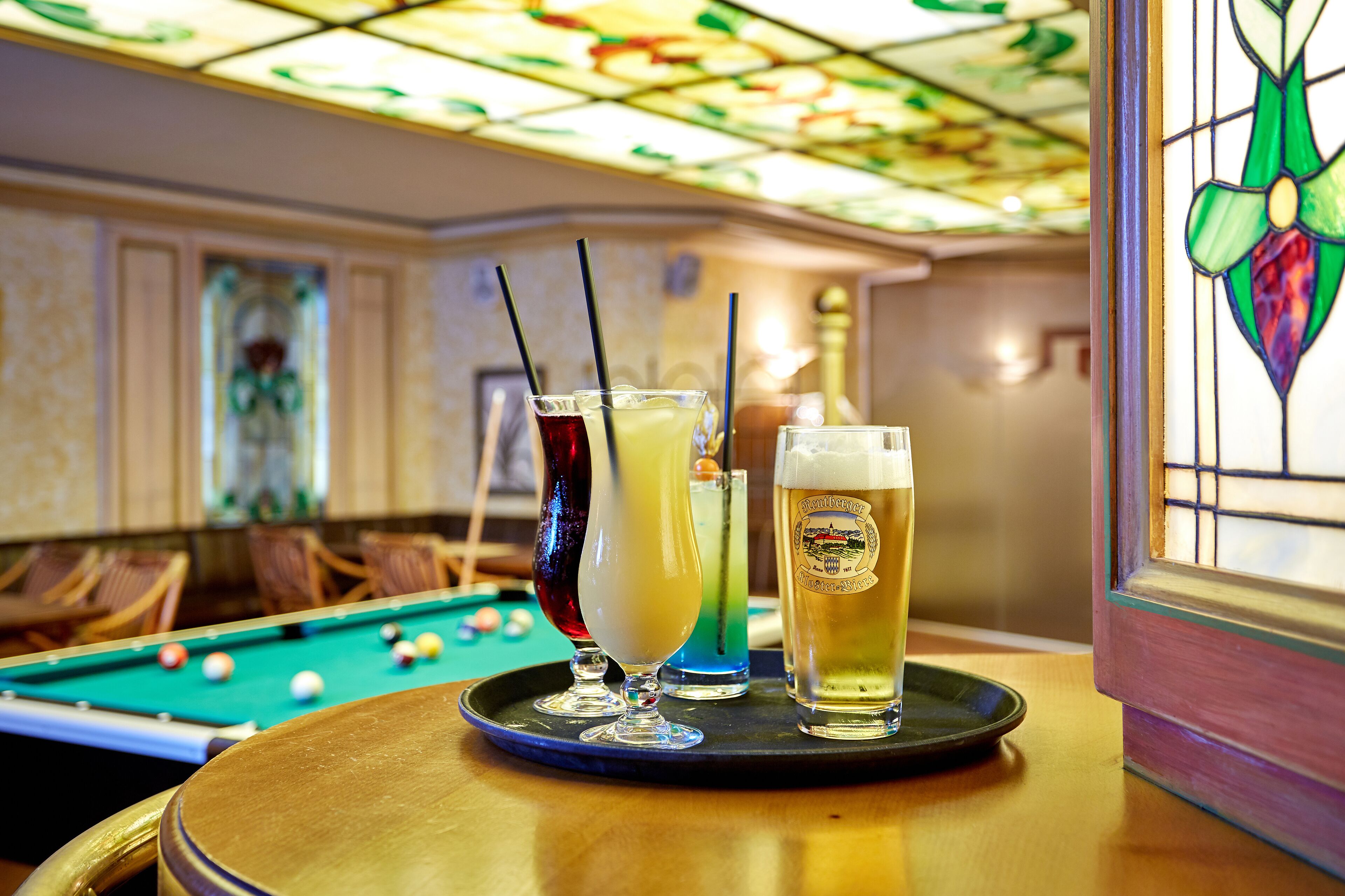 A pool table with balls, a bar area with glasses of beer, and a stained glass window.