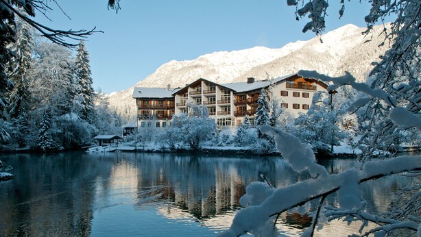 Un edificio cubierto de nieve con múltiples balcones que dan a un lago tranquilo, rodeado de árboles cargados de nieve y montañas al fondo.