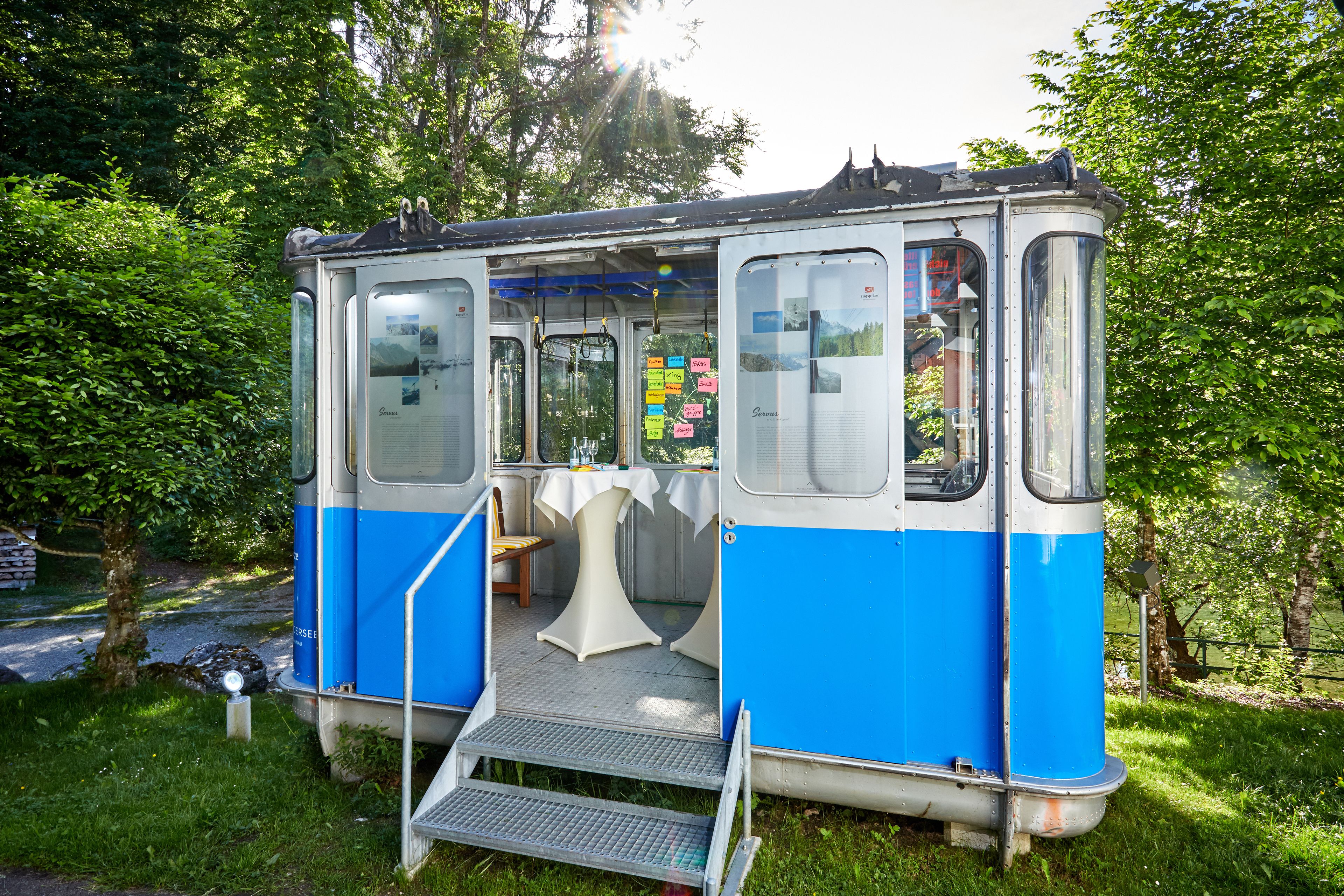 A small, enclosed bus with a table and chairs inside, surrounded by greenery.