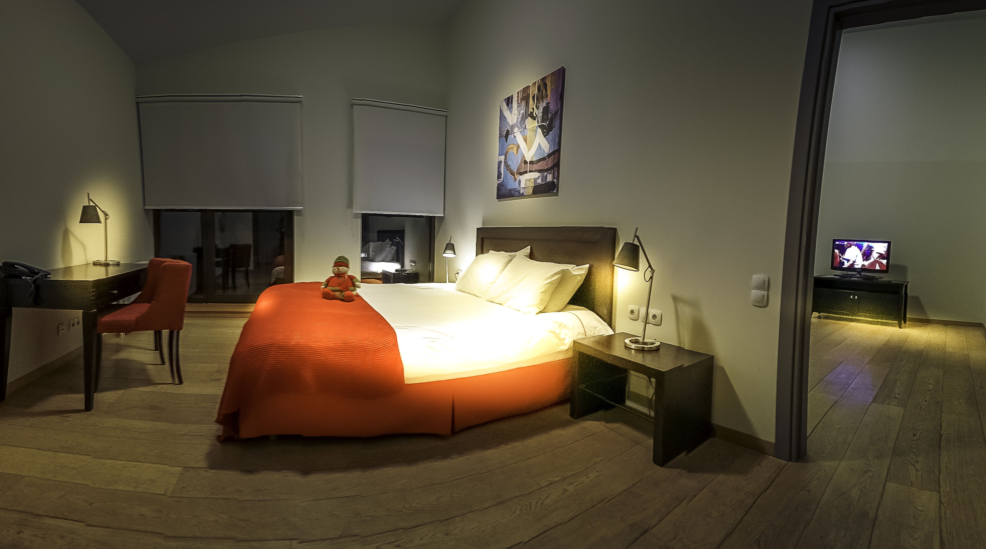 A modern hotel room with a large bed, a desk with a chair, a TV, and a painting on the wall.