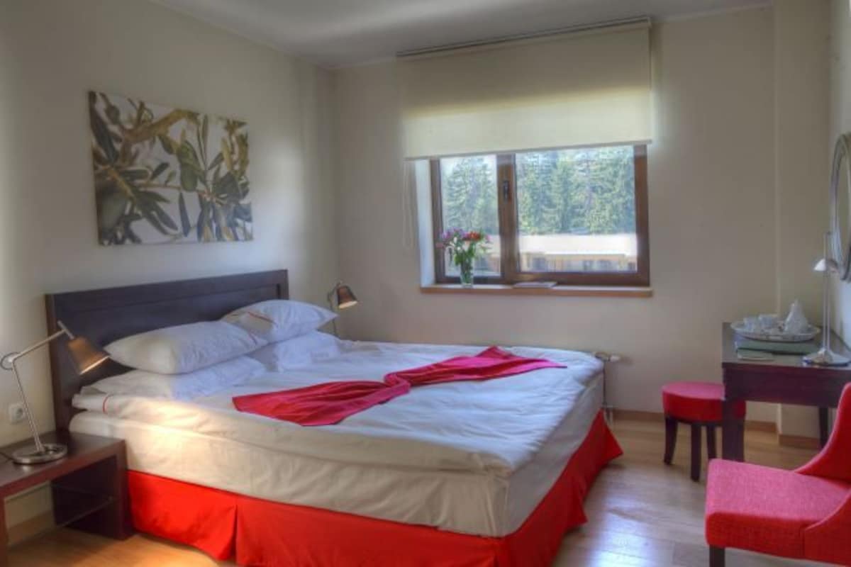 A hotel room with a large bed, a desk, a chair, a window with a view of trees, and a painting on the wall.
