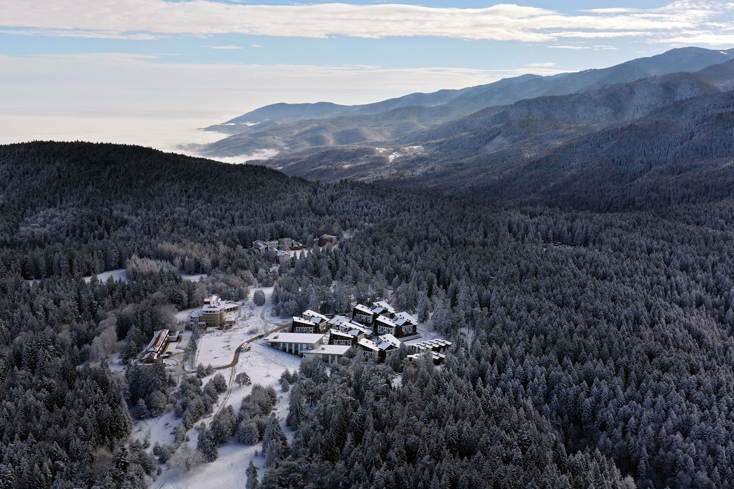 Un refugio cubierto de nieve con varias construcciones en medio de un bosque frondoso, rodeado de montañas.