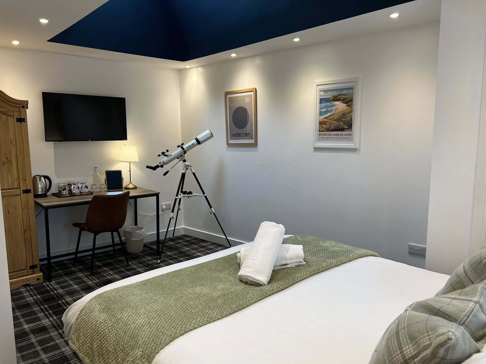 Et hotelværelse med en seng, et skrivebord med et teleskop, et fjernsyn og to billeder hængende på væggen.