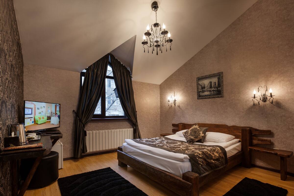 A bedroom with a bed, a television, a radiator, a window with curtains, and a chandelier.