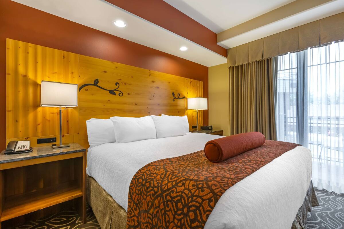A hotel room with a large bed, wooden headboard, two bedside lamps, a telephone, and a window with curtains.