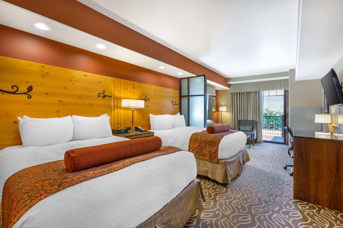 A hotel room with two beds, a desk, a chair, and a balcony with a view.