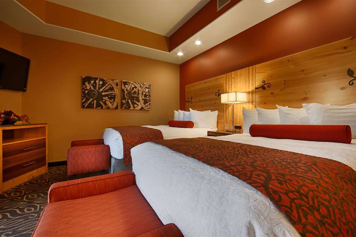A hotel room with two beds, a wooden headboard, a desk with a lamp, and artwork on the wall.