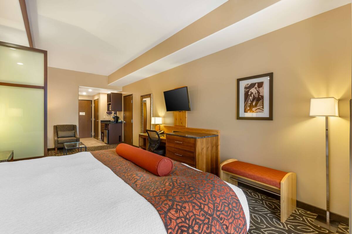 A hotel room with a large bed, a desk with a chair, a television, and a small bench.
