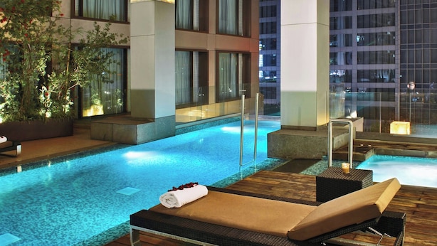 Un área de piscina en la azotea con sillones de descanso y un jacuzzi pequeño, rodeada de edificios modernos.