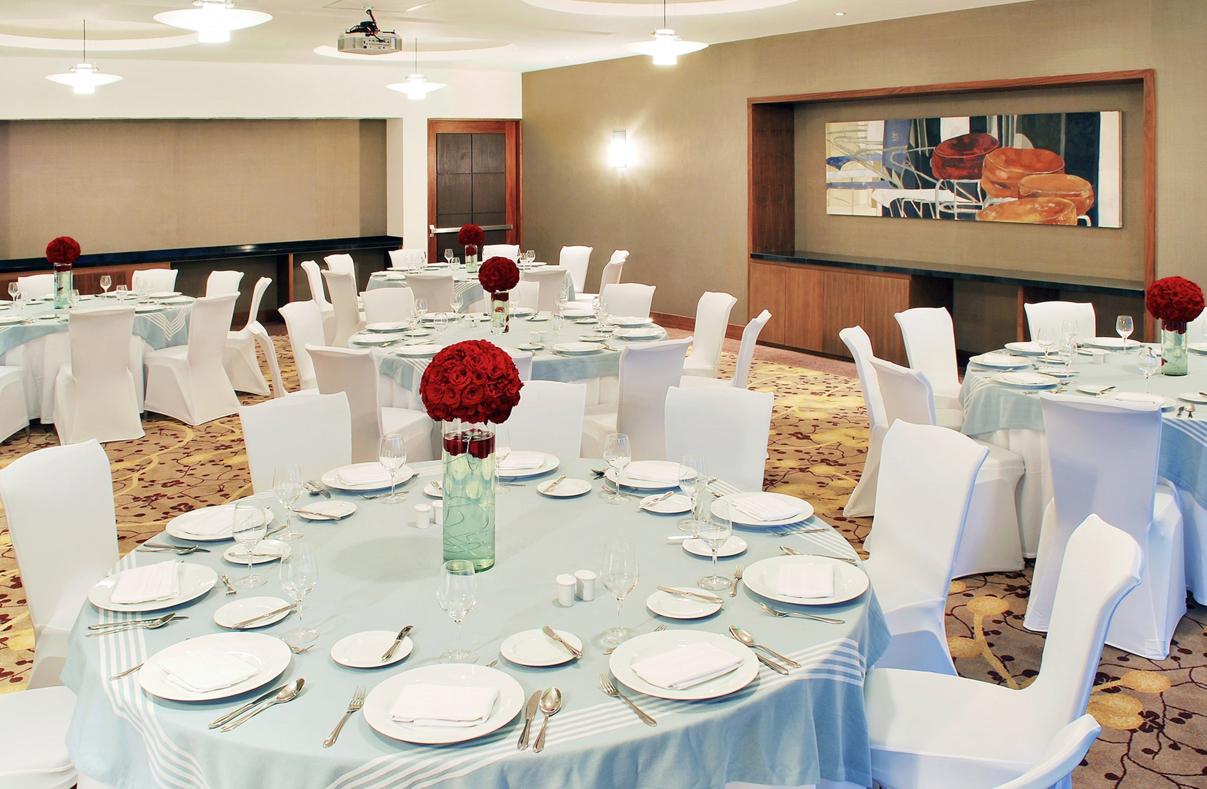 Uma sala de banquetes com mesas redondas preparadas para refeições, cadeiras brancas e uma tela de projeção na parede.