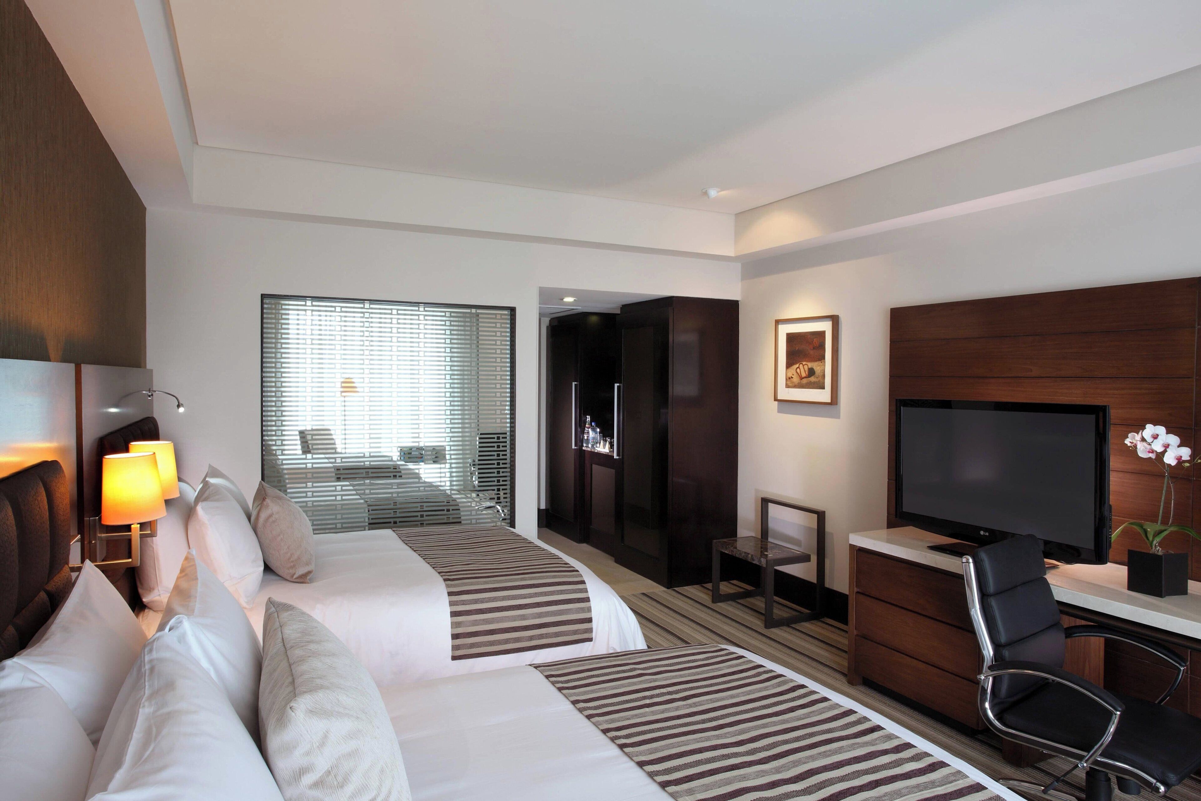 A hotel room with two beds, a desk with a TV, a chair, and a window with blinds.