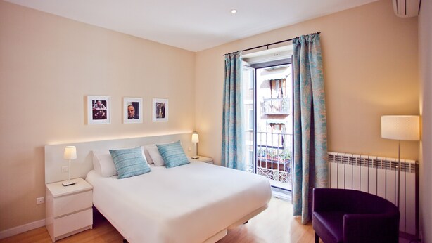Habitación de hotel con una cama grande, una mesita de noche, una silla y vistas a un balcón a través de una ventana con cortinas.