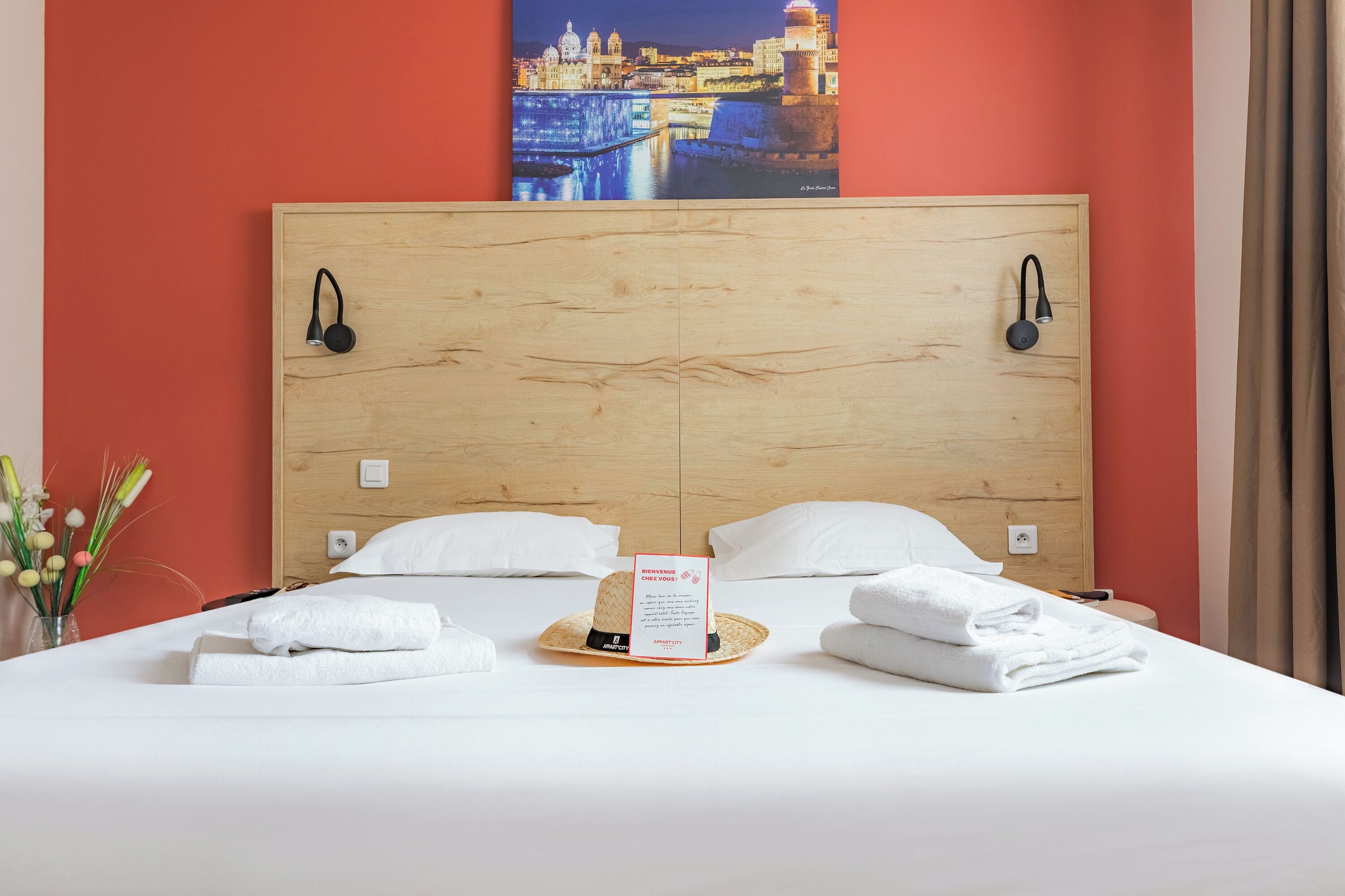 Habitación de hotel con cabecera de madera, dos lámparas de noche, cama con ropa blanca, una tarjeta de bienvenida y un adorno en la pared.