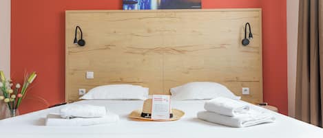 Habitación de hotel con cabecera de madera, dos lámparas de noche, cama con ropa blanca, una tarjeta de bienvenida y un adorno en la pared.