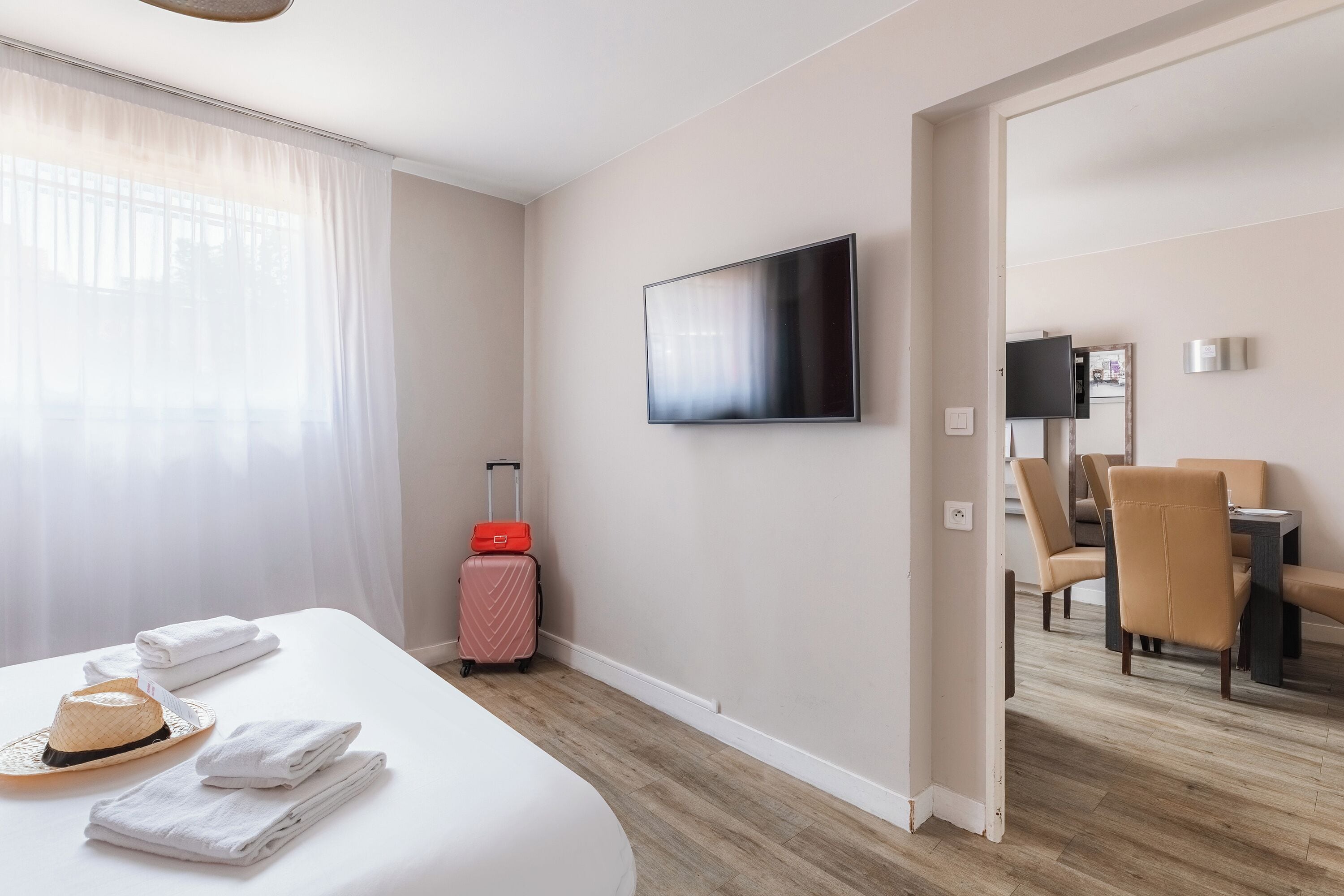 A hotel room with a bed, a TV, a dining table with chairs, and a suitcase.