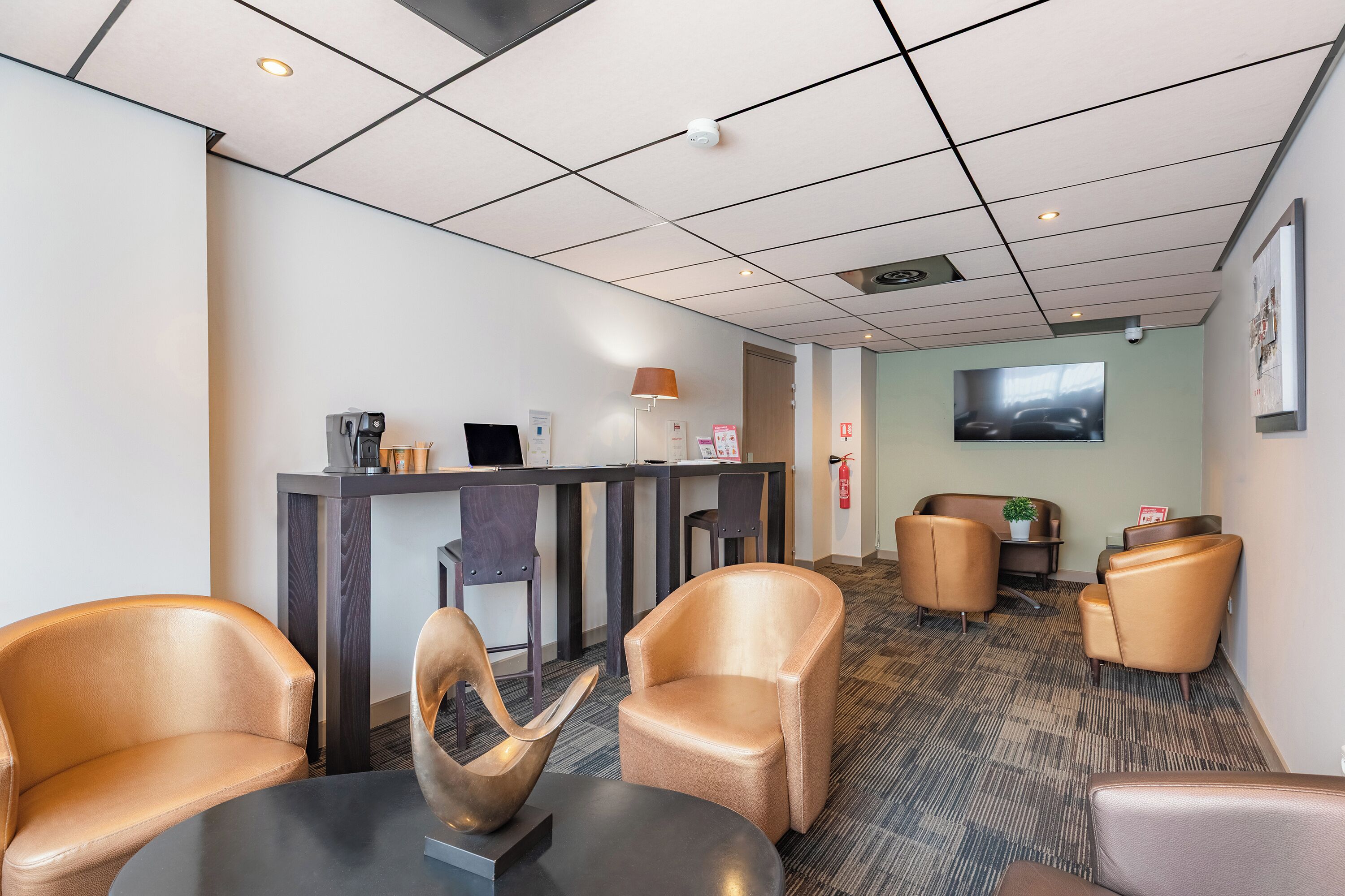 Een moderne hotel lobby met een receptiebalie, lederen fauteuils, een koffieapparaat en een flatscreen televisie aan de muur.