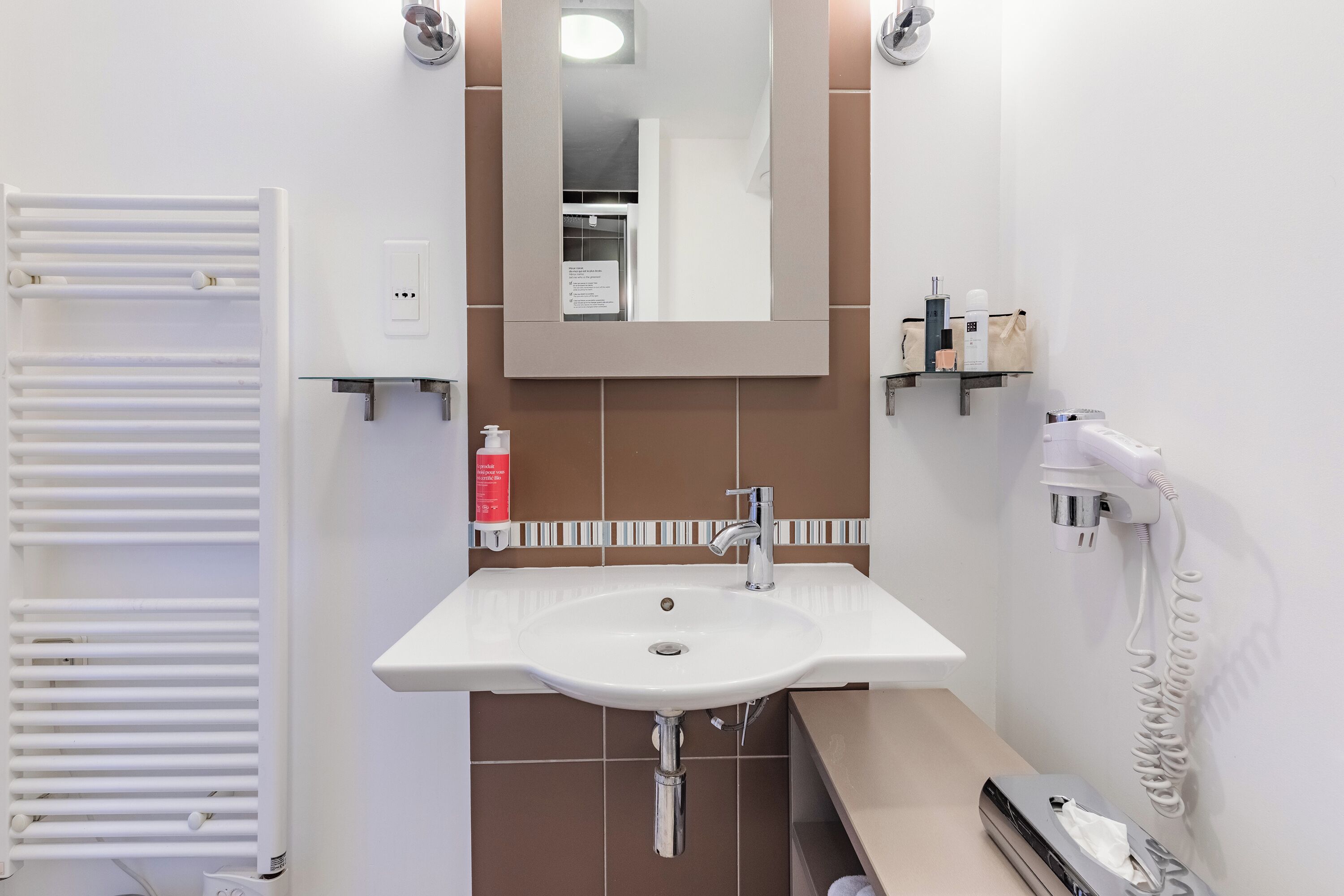 Une salle de bain moderne avec un lavabo blanc, un miroir, un porte-serviettes et un sèche-cheveux posés sur une étagère fixée au mur.