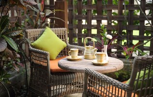 A wicker chair with a green cushion, a small round table with a tea set, and a wooden fence in the background.