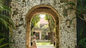 Un arc en pierre mène à une cour dallée, entourée d’une végétation luxuriante et bordée, au fond, d’un bâtiment.